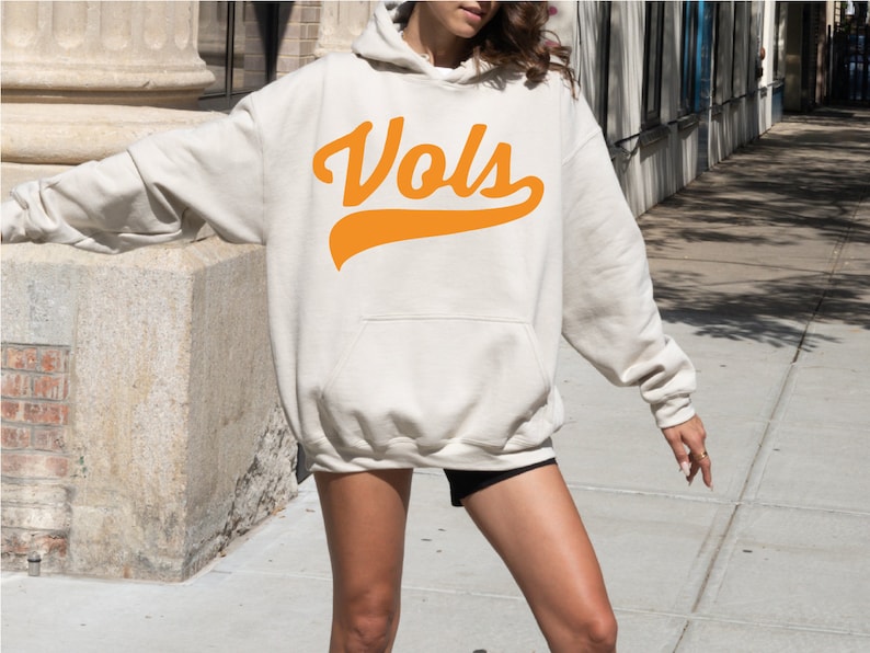 May include: An oversized cream-colored hooded sweatshirt featuring a large orange graphic on the chest with the text "Vols" in a cursive script font. The garment includes long sleeves, a front kangaroo pocket, and a drawstring hood.