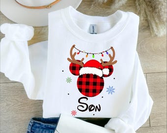 Puede incluir: Sudadera blanca de manga larga con una cabeza de Mickey Mouse de cuadros rojos y negros que lleva un gorro de Papá Noel y cuernos de reno. La sudadera tiene la palabra "Hijo" escrita en una fuente de Disney debajo de la cabeza de Mickey Mouse.