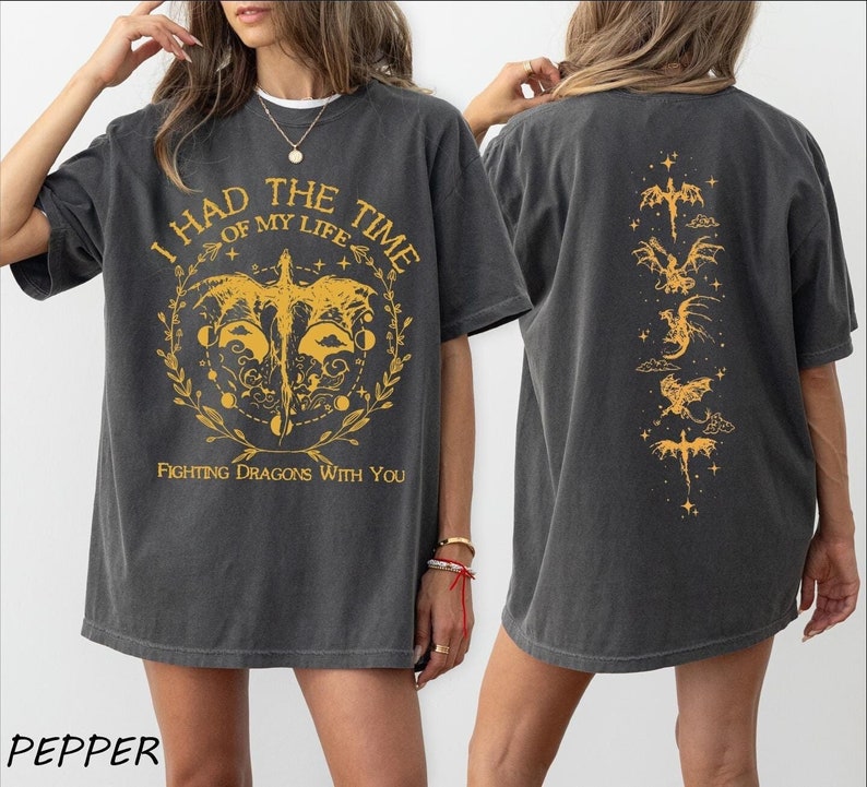 Puede incluir: Una camiseta negra de gran tama&ntilde;o con un estampado gr&aacute;fico amarillo en la parte delantera que dice "Tuve el mejor momento de mi vida luchando contra dragones contigo". La parte posterior de la camiseta tiene una fila vertical de siluetas de dragones amarillos.