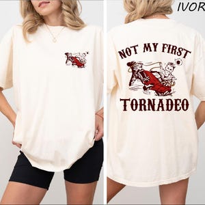 May include: A white t-shirt with a red graphic of a person riding a red car in a tornado. The text "NOT MY FIRST TORNADEO" is printed in red on the back of the shirt.