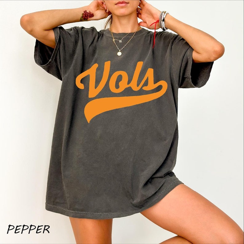 May include: A charcoal gray short-sleeved t-shirt featuring the word Vols printed in a bold orange script font across the chest. The garment has a crew neckline and a relaxed fit, with the color name Pepper printed in the bottom left corner.