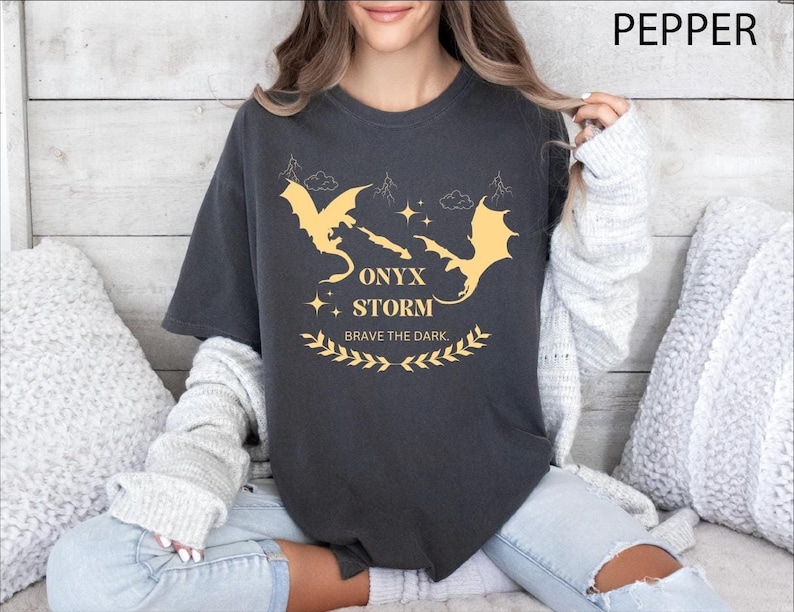 May include: A dark grey t-shirt with a graphic of two gold dragons and the text "ONYX STORM" and "BRAVE THE DARK".