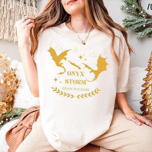 May include: A white t-shirt featuring a gold graphic of two dragons facing each other. The text "ONYX STORM" and "BRAVE THE DARK." is printed below the dragons.