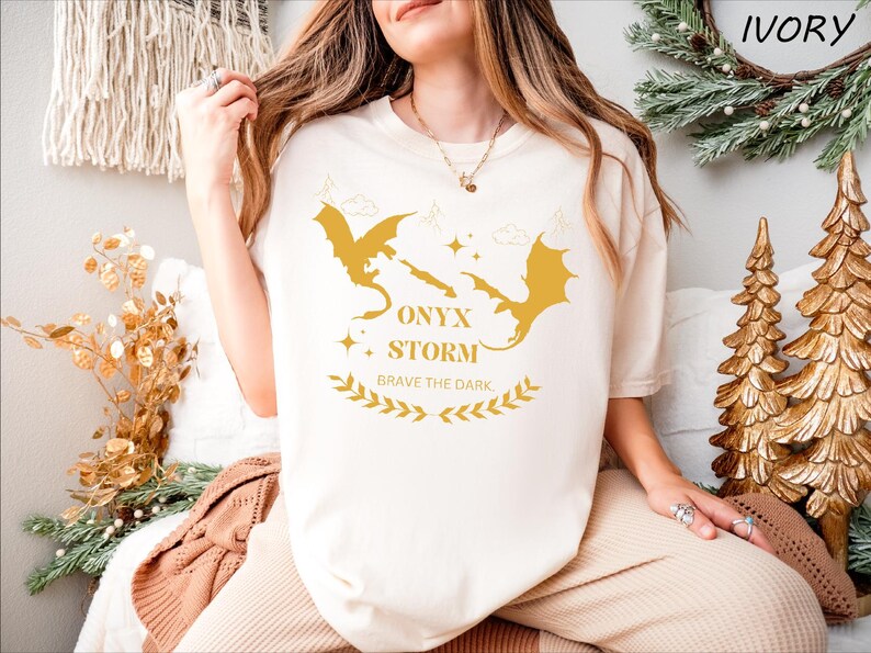 May include: A white t-shirt featuring a gold graphic of two dragons facing each other. The text "ONYX STORM" and "BRAVE THE DARK." is printed below the dragons.
