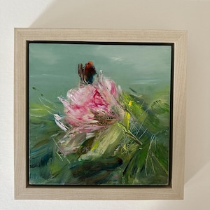 May include: An original oil painting of a pink flower with a butterfly, set against a green background. The artwork is framed in a light-colored wooden frame. The painting features visible brushstrokes and a textured appearance.