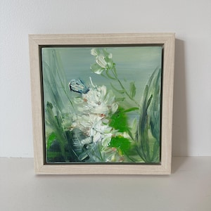 May include: Square framed oil painting of white flowers and green foliage against a soft green background. A small blue butterfly rests on the flowers. The artwork is in a light-colored wooden frame, creating a serene and natural aesthetic.