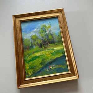 Peut inclure: Peinture de paysage encadr&eacute;e dans un cadre dor&eacute;. L'&oelig;uvre repr&eacute;sente un champ vert vibrant et des arbres sous un ciel bleu nuageux. La peinture utilise des coups de pinceau visibles pour cr&eacute;er de la texture et de la profondeur.