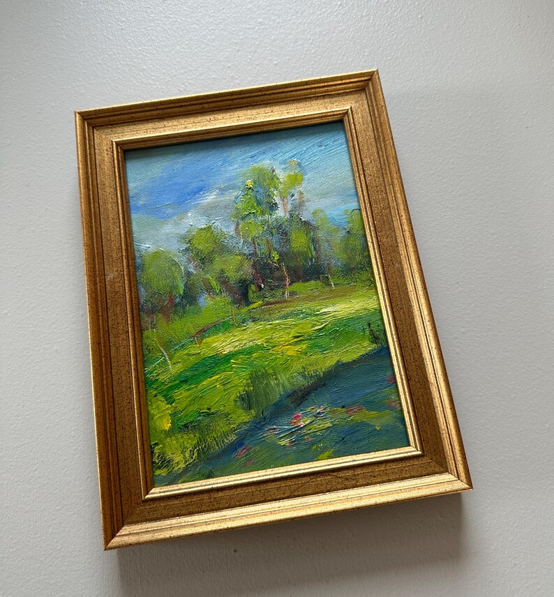 Peut inclure: Peinture de paysage encadr&eacute;e dans un cadre dor&eacute;. L'&oelig;uvre repr&eacute;sente un champ vert vibrant et des arbres sous un ciel bleu nuageux. La peinture utilise des coups de pinceau visibles pour cr&eacute;er de la texture et de la profondeur.