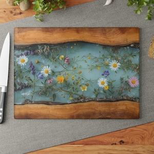 Könnte beinhalten: Ein rechteckiges Holzschneidebrett mit einem hellblauen Harzmittelteil, eingelegt mit bunten Wildblumen. Das Brett hat einen natürlichen Holzmaserungsrand und wird von einem Edelstahlmesser begleitet.