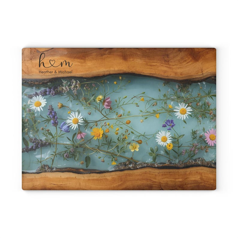 Puede incluir: Tabla de servir rectangular de madera con un centro de resina azul claro, con incrustaciones de flores silvestres de colores. La tabla tiene un borde de vetas de madera natural y el texto "h♡m Heather & Michael" en la esquina superior izquierda.