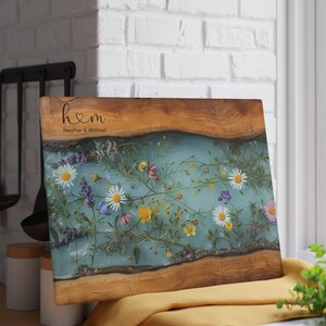 Puede incluir: Tabla de cortar rectangular de madera con un centro de resina azul claro, con flores silvestres de colores incrustadas. La tabla tiene un borde de madera natural y el texto "h♡m Heather & Michael" est&aacute; grabado.