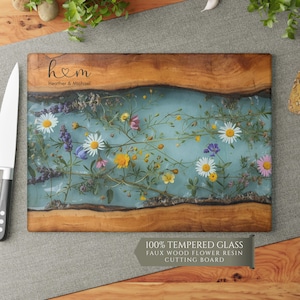 Puede incluir: Tabla de cortar rectangular con un centro de resina azul claro, con incrustaciones de flores silvestres de colores. La tabla tiene un borde de madera natural y el texto "h♡m Heather & Michael". La esquina inferior derecha tiene una etiqueta que dice "100% TEMPERED GLASS FAUX WOOD FLOWER RESIN CUTTING BOARD".