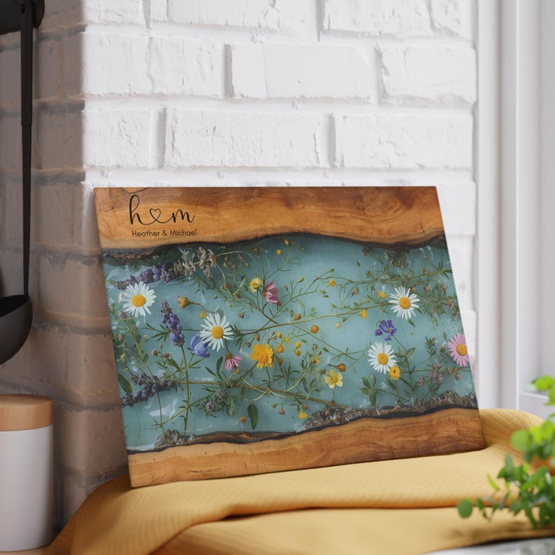 Puede incluir: Tabla de cortar rectangular de madera con un centro de resina azul claro, incrustada con flores silvestres de colores. La tabla tiene un borde de madera natural y el texto "h♡m Heather & Michael" est&aacute; grabado en la parte superior.