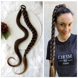 Peut inclure: Une longue extension de cheveux tressée brune avec un élastique noir. L'extension est présentée sur une surface blanche et également portée par une personne avec un t-shirt noir qui dit "MERCI -98- PARIS".