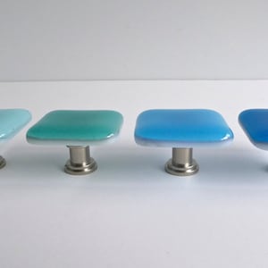 Peut inclure: Quatre boutons de cabinet carrés en verre avec une base blanche et un dégradé de couleurs bleu, turquoise et bleu clair. Les boutons ont une base en métal argenté.