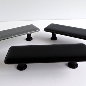 May include: Three rectangular cabinet pulls. One is grey, and two are black. Each pull has a rounded rectangular shape with a glossy finish and is mounted on dark brown cylindrical posts.