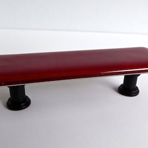 May include: A red glass cabinet pull with black metal hardware. The pull is rectangular and has rounded edges.