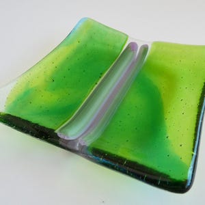 May include: A handcrafted glass dish with a square shape and curved edges. The dish features shades of green and a central stripe with purple and white hues. The glass has a textured appearance.