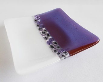 May include: A small, square glass dish with a white and purple design. The dish has a border of small purple flowers with black centers.