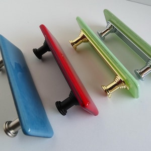 May include: Four rectangular glass cabinet knobs with different colored glass and metal hardware. The knobs are in a row from left to right: light blue, red, yellow, and green.