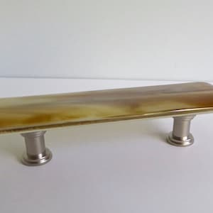 May include: A rectangular cabinet pull with a marbled, amber-coloured glass top. The glass is mounted on two brushed nickel posts. Suitable for drawers and cabinets. The length is approximately 15 cm.