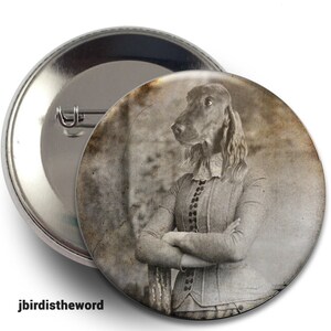 May include: A vintage-style button featuring a sepia-toned image of a dog wearing a dress and standing with its arms crossed. The dog has a serious expression and is looking to the side. The background is blurred and appears to be a forest.