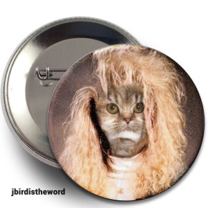 May include: A round button pin featuring a cat with a blonde wig. The cat has green eyes and a serious expression. The pin is silver and has a safety pin back. The text "jbirdistheword" is at the bottom.