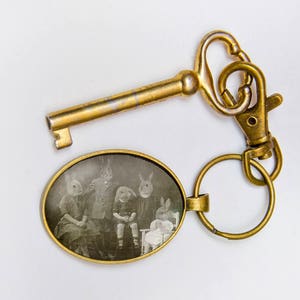 May include: A brass key chain with a key and a round, oval-shaped charm. The charm has a black and white photo of four people wearing rabbit masks.