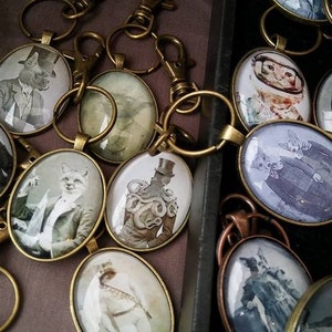 May include: A collection of keyrings featuring various animal illustrations in oval-shaped glass medallions. The medallions are attached to brass key rings and swivel clasps. The illustrations include a fox, a dog, a cat, and a creature with an octopus-like body.