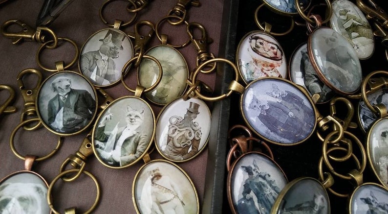 May include: A collection of keyrings featuring various animal illustrations in oval-shaped glass medallions. The medallions are attached to brass key rings and swivel clasps. The illustrations include a fox, a dog, a cat, and a creature with an octopus-like body.