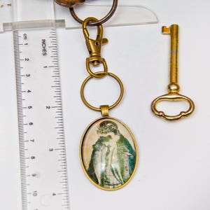 May include: A gold-toned key chain with a round, glass charm featuring a vintage image of a woman in a hat. The charm is attached to a gold-toned key ring and a gold-toned lobster clasp. A separate gold-toned skeleton key is also included.