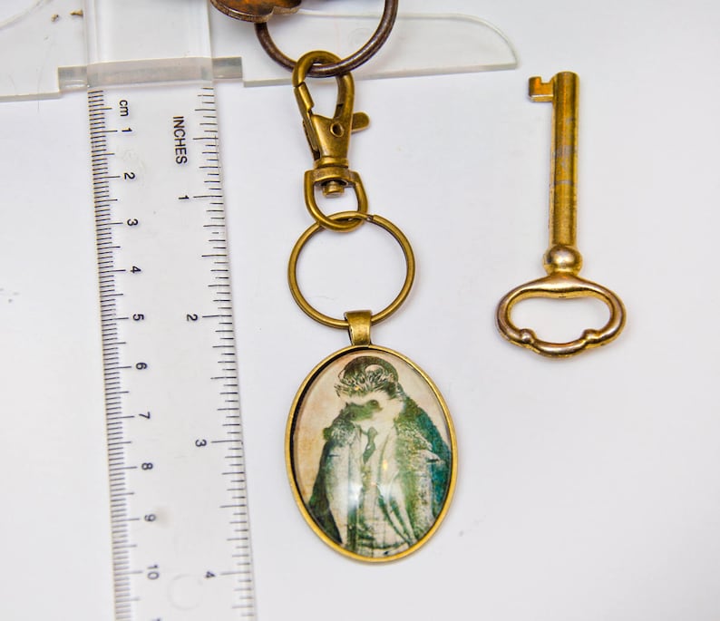 May include: A gold-toned key chain with a round, glass charm featuring a vintage image of a woman in a hat. The charm is attached to a gold-toned key ring and a gold-toned lobster clasp. A separate gold-toned skeleton key is also included.