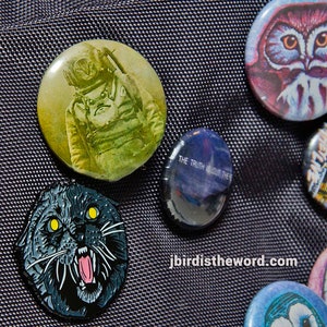 May include: A collection of nine colourful button badges featuring various designs, including an astronaut, an owl, a cat, and a galaxy. The badges are arranged on a black fabric background.