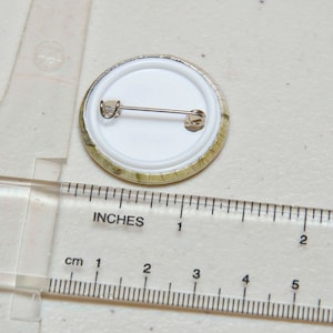May include: A round button with a white background and a gold rim. The button has a pin back and is approximately 2.5 cm in diameter.