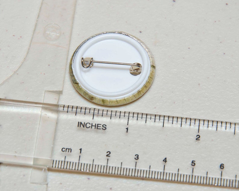 May include: A round button with a white background and a gold rim. The button has a pin back and is approximately 2.5 cm in diameter.