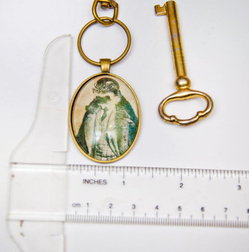 May include: A brass key chain with a round, oval-shaped charm featuring a vintage image of a person wearing a hat and coat. The charm is attached to a brass key ring. A separate brass skeleton key is also included.