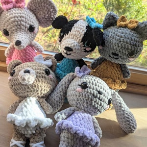 May include: Five crocheted stuffed animals in various colors and styles. A gray mouse with pink ears and a pink bow, a black and white dog with blue bow, a gray cat with brown bow, a brown bear with a white dress, and a gray bunny with a purple dress.
