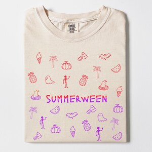 May include: A folded beige t-shirt with a crew neck. The shirt features red and purple doodles of ice cream cones, palm trees, pumpkins, ghosts, and the word "SUMMERWEEN". The shirt is made by Comfort Colors.
