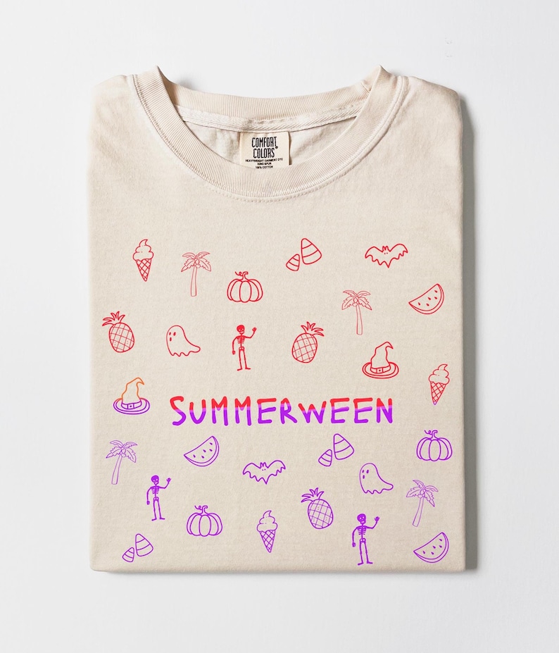 May include: A folded beige t-shirt with a crew neck. The shirt features red and purple doodles of ice cream cones, palm trees, pumpkins, ghosts, and the word "SUMMERWEEN". The shirt is made by Comfort Colors.