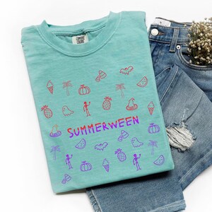 May include: A turquoise t-shirt with a colorful graphic design featuring Halloween and summer-themed icons. The word "SUMMERWEEN" is printed in purple and red. The shirt is paired with blue denim jeans.