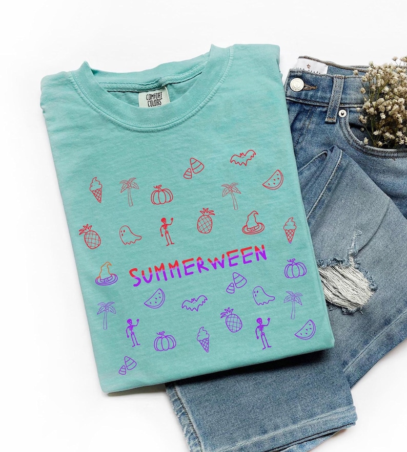 May include: A turquoise t-shirt with a colorful graphic design featuring Halloween and summer-themed icons. The word "SUMMERWEEN" is printed in purple and red. The shirt is paired with blue denim jeans.