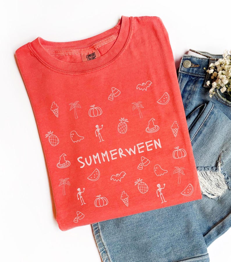 May include: Coral-colored t-shirt with white graphic icons and the word "SUMMERWEEN". The design includes Halloween and summer-themed illustrations like pumpkins, bats, and ice cream cones. The shirt is paired with blue denim jeans.
