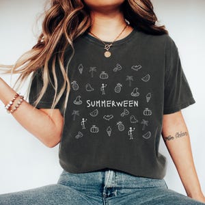 May include: Dark gray t-shirt with the word "SUMMERWEEN" in white, surrounded by white doodle illustrations of ice cream cones, palm trees, pumpkins, candy corn, bats, and ghosts. The shirt is a relaxed fit.