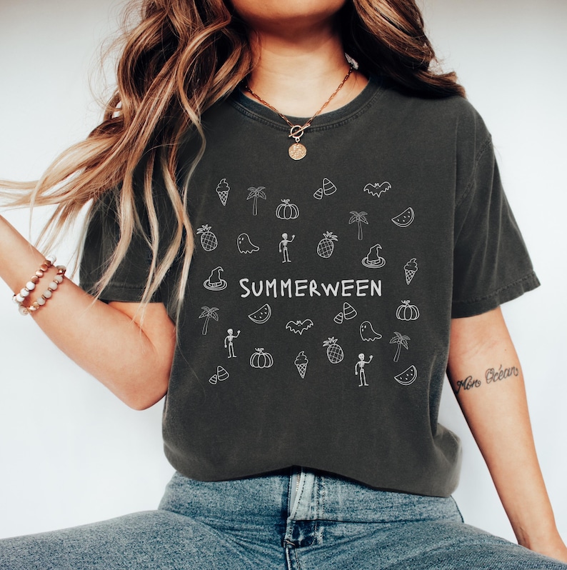 May include: Dark gray t-shirt with the word "SUMMERWEEN" in white, surrounded by white doodle illustrations of ice cream cones, palm trees, pumpkins, candy corn, bats, and ghosts. The shirt is a relaxed fit.