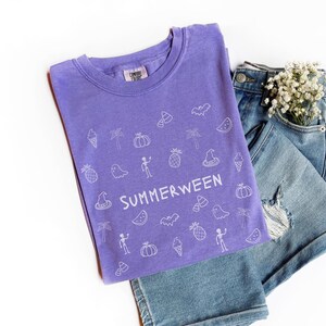 May include: A purple t-shirt with the word "SUMMERWEEN" and various white illustrations of Halloween and summer-themed objects. The shirt is paired with denim shorts and a small bouquet of white flowers.