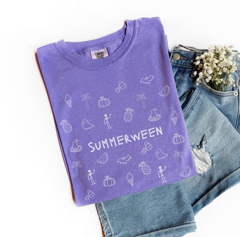 May include: A purple t-shirt with the word "SUMMERWEEN" and various white illustrations of Halloween and summer-themed objects. The shirt is paired with denim shorts and a small bouquet of white flowers.