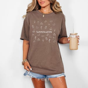 May include: Brown t-shirt with the word "SUMMERWEEN" and various summer and Halloween-themed doodles in white. The shirt is paired with denim shorts. A glass jar with a straw is held in the hand.