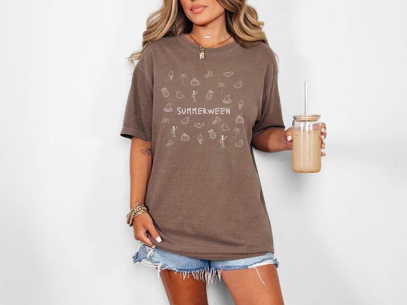 May include: Brown t-shirt with the word "SUMMERWEEN" and various summer and Halloween-themed doodles in white. The shirt is paired with denim shorts. A glass jar with a straw is held in the hand.