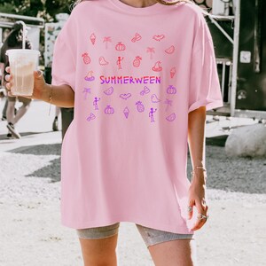 May include: A light pink t-shirt with the word "SUMMERWEEN" in a gradient of red and purple. The shirt features a pattern of Halloween and summer-themed doodles in red and purple.
