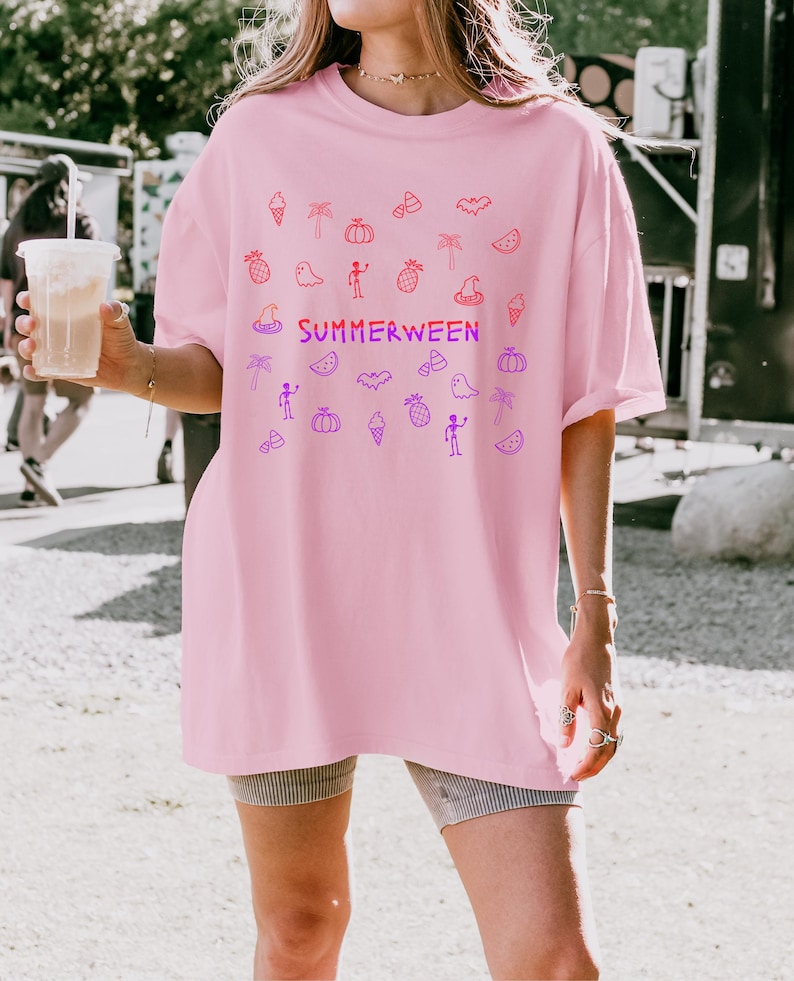 May include: A light pink t-shirt with the word "SUMMERWEEN" in a gradient of red and purple. The shirt features a pattern of Halloween and summer-themed doodles in red and purple.
